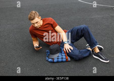 Ein hübscher, modischer Studententyp in stylischer Freizeitkleidung mit Jeans liegt auf dem Asphalt und benutzt ein Smartphone Stockfoto