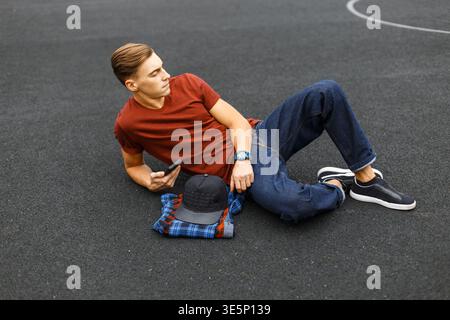 Stilvoller, hübscher junger Mann in modischer Kleidung mit T-Shirt und Jeans liegt mit einem Smartphone auf dem Asphalt des Stadions Stockfoto