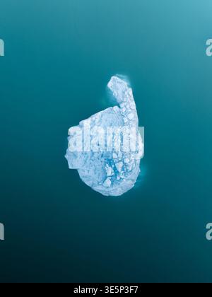Aus der Vogelperspektive eines einsamen, geformten Eisbergs, der sich in der Gletscherlagune befindet, dessen eisblaue Form im Kontrast zum dunklen, ruhigen Wasser, Jokulsarlon, Eis steht Stockfoto