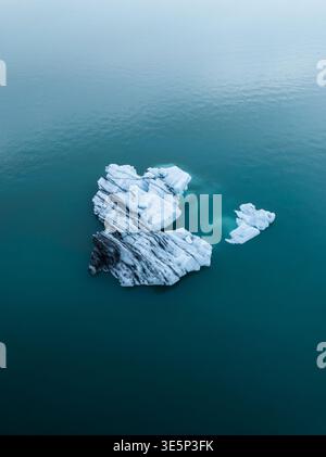 Aus der Vogelperspektive zieht sich ein einsamer Eisberg in der Weite der Lagune, dessen eiskaltes Blau im Kontrast zum tiefen türkisfarbenen Wasser steht, Jokulsarlon, Sveitarfélagið Stockfoto