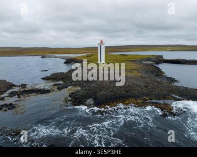 Aus der Vogelperspektive eines scharf weißen Leuchtturms mit einer roten Spitze, die hoch auf einem Felsvorsprung vor dem stimmungsvollen Himmel steht, Kálfshamarsvík, Húnabyggð, Island. Stockfoto