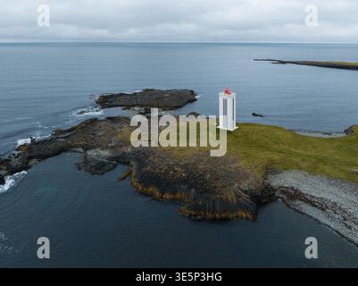Aus der Vogelperspektive eines weißen Leuchtturms mit einem roten Dach steht Wache auf einer zerklüfteten, grünen Insel vor dem steinigen, grauen Meer, Kálfshamarsvík, Stockfoto