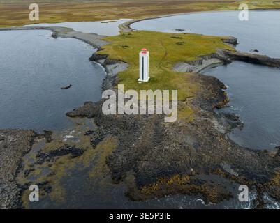 Aus der Vogelperspektive eines einsamen weißen Leuchtturms mit einer roten Spitze stehenden Wache auf einem Felsvorsprung inmitten des dunklen, wirbelnden Wassers, Kálfshamarsvík, Island Stockfoto