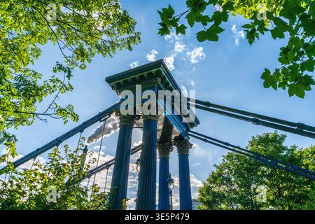 Ein flacher Blick auf die klassischen Säulen und Kabel einer Hängebrücke, eingerahmt von grünen Bäumen und hellblauem Himmel, kombiniert urbane Architektur und Motor Stockfoto