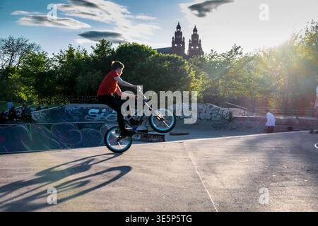 Ein junger Fahrer führt einen BMX-Wheelie durch einen mit Graffiti bedeckten Skatepark in der Nähe der Kelvingrove Art Gallery in Glasgow aus, der von der späten Sonne beleuchtet und umgeben ist Stockfoto