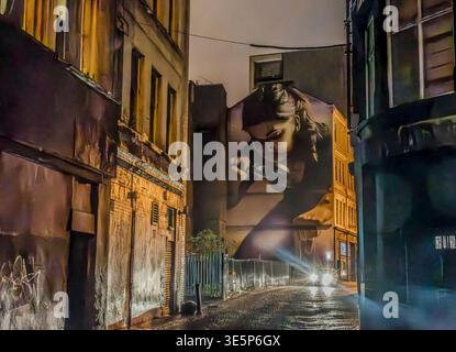 Ein großes fotorealistisches Wandbild eines Mädchens mit einer Lupe füllt nachts eine enge nasse Gasse, eingerahmt von verwitterten Gebäuden, Graffiti und Bright Stockfoto
