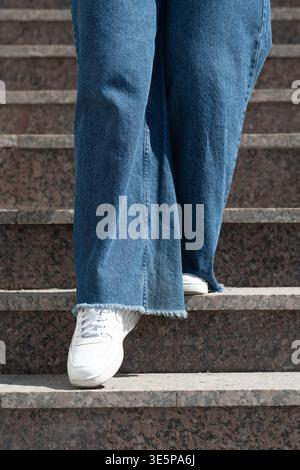 Niedriger Blickwinkel auf eine Person mit breiten Beinen in blauer Jeans und weißen Turnschuhen, die die Granittreppen hinuntergeht. Stockfoto