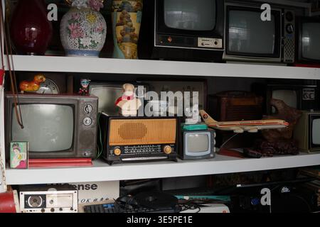 Regale mit vielen Retro-Fernsehern, einem alten Radio und antiken Gegenständen schaffen eine nostalgische Szene mit traditionellen asiatischen Vasen und historischen Elementen Stockfoto