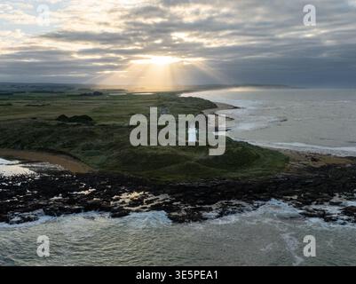 Aus der Vogelperspektive auf den Leuchtturm, der bewacht wird, während die Sonne durch die Wolken über der zerklüfteten Küste in Otara, Southland Region, Neuseeland eindringt. Stockfoto