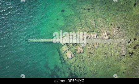 Aus der Vogelperspektive auf die zerklüftete Küste trifft auf das türkisfarbene Meer, wo sich ein Steinpier erstreckt, ein starker Kontrast zum klaren Wasser, Carantec, Stockfoto