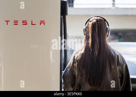 Frankfurt, Deutschland. Eine Person mit Kopfhörern steht neben der Tesla-Ladestation und zeigt den alltäglichen Gebrauch von Elektrofahrzeugen. Stockfoto