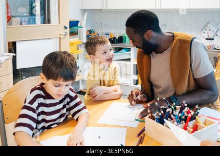 Reifer männlicher Lehrer sitzt mit zwei Schülern am Tisch während des Kunstunterrichts in der Vorschule Stockfoto