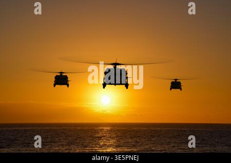 Militärhubschrauber fliegen in Formation bei Sonnenuntergang über dem Meer unter militärischen Konfliktbedingungen. Stockfoto