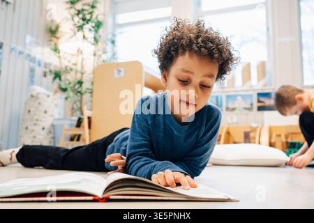 Junge mit lockigen Haaren liest Buch, während er auf dem Boden des Klassenzimmers liegt Stockfoto