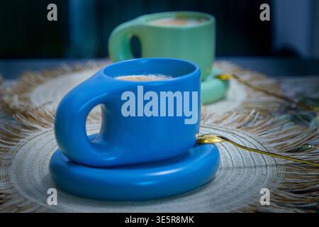 Latte serviert in einem modernen blauen Designer-Becher auf einem rustikalen, gewebten Tischset mit einer zweiten Tasse, die im Hintergrund weich verwischt ist, gemütliche Stillleben-Szene Stockfoto
