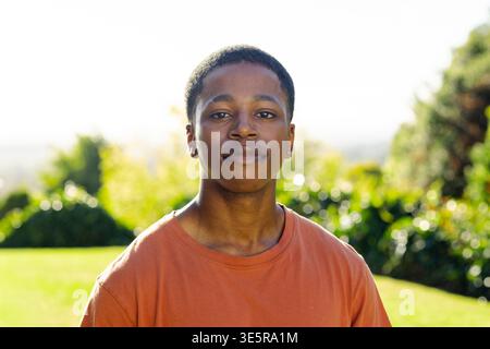Afroamerikanischer Mann in Zwanzigern, der auf dem sonnendurchfluteten Rasen stand, mit orangefarbenem T-Shirt und Creolen Stockfoto
