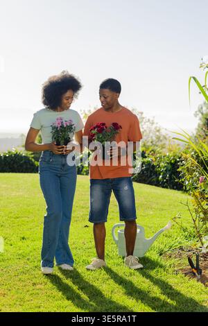 Afroamerikanisches Paar, das im Hinterhof steht und rosafarbene und rote Blumen mit einer Gießkanne hält Stockfoto