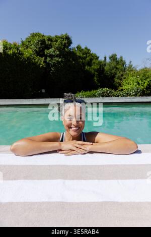 Afroamerikanische Frau, die zu Hause am Pool ausruht, sich auf Handtuch am Rand in Badeanzug, Sonnenbrille lächelnd Stockfoto