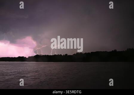 Mehrere Blitze treffen über einen dunklen Wald hinweg, beleuchten Sturmwolken und reflektieren nachts schwach auf der Seenoberfläche. Stockfoto