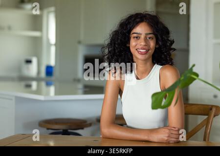 Afroamerikanerin sitzt an einem Holztisch in der modernen Küche und lächelt mit Pflanzenblatt in die Kamera Stockfoto