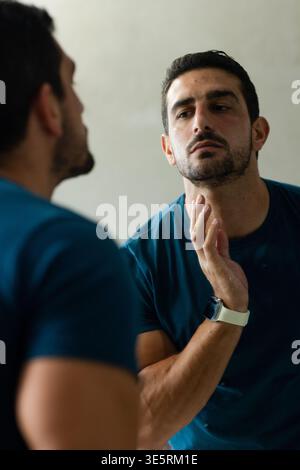 Mann, der den Kiefer berührt, die Reflexion im Wandspiegel überprüft, weiße Smartwatch und blaues T-Stück trägt Stockfoto