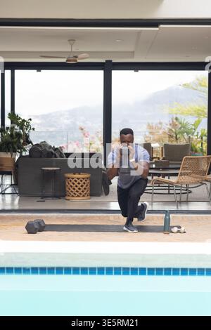 Kettlebell und Hexadezimal Kurzhanteln liegen auf einer schwarzen Matte am Pool, Schiebetüren zeigen ein Sofa Stockfoto