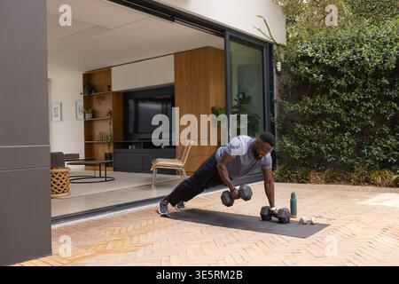 Afroamerikanischer Mann mit grauem Hemd, der auf einer Terrassenmatte trainiert und Hexenhanteln hält, Kopierraum Stockfoto