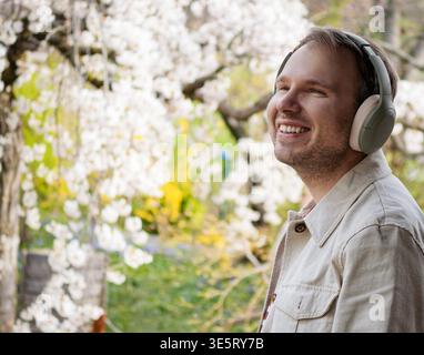 Ein lächelnder Mann mit Kopfhörern genießt Musik zwischen blühenden Frühlingsbäumen in einem japanischen Park und schafft so eine filmische Kulisse. Stockfoto