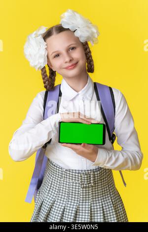 Fröhliches Schulmädchen mit Zöpfen und Rucksack hält das Smartphone mit grünem Chroma-Schlüsselbildschirm. Mädchen lächelt vor hellgelbem Hintergrund. Ideal für Stockfoto