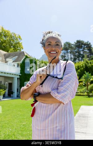Frau mittleren Alters mit gestreiftem Kleid und Quastengürtel, die auf dem Vorhof steht und eine dunkle Sonnenbrille hält Stockfoto