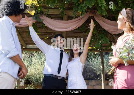 Frisch vermähltes Paar hebt die Arme, lacht unter der Pergola, Braut im Spitzenkleid, Bräutigam in weißem Hemd Stockfoto