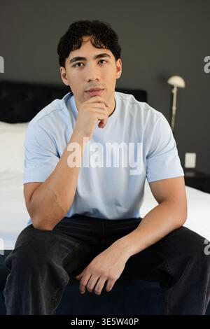 Asiatischer Mann sitzt auf Fußbrett im Schlafzimmer, trägt hellblaues T-Stück und dunkle Jeans, neben der Lampe Stockfoto