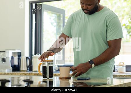 Afroamerikaner, der in der Küche Kaffee macht, mit französischer Presse, mintfarbenem T-Shirt, Kopierraum Stockfoto