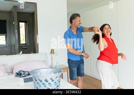 Ein älteres Paar tanzt spielerisch neben dem Bett im Schlafzimmer mit Wäschekorb und Nachttischlampe Stockfoto