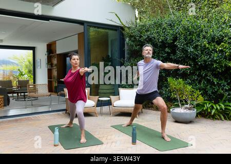 Seniorenpaar üben Yoga im Stehen in Sportbekleidung auf der Terrasse mit Matten und Wasserflaschen Stockfoto
