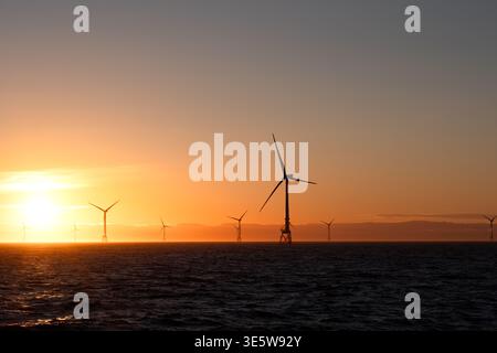 Eine Reihe von Offshore-Windturbinen, die sich vor einem leuchtenden orange-goldenen Sonnenuntergang über der Nordsee abzeichnen, mit sichtbaren Hügeln im Hintergrund. Stockfoto