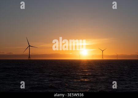 Eine Reihe von Offshore-Windturbinen, die sich vor einem leuchtenden orange-goldenen Sonnenuntergang über der Nordsee abzeichnen, mit sichtbaren Hügeln im Hintergrund. Stockfoto