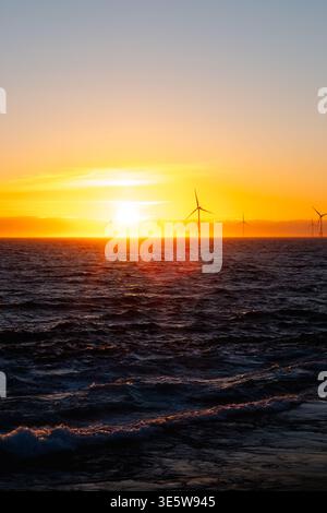 Eine Reihe von Offshore-Windturbinen, die sich vor einem leuchtenden orange-goldenen Sonnenuntergang über der Nordsee abzeichnen, mit sichtbaren Hügeln im Hintergrund. Stockfoto