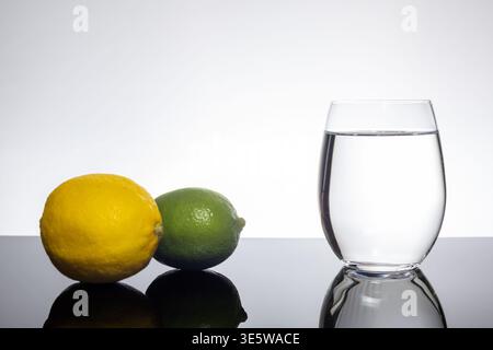 Zitrone und Limette mit Glas Wasser auf schwarzer reflektierender Oberfläche mit weißem Hintergrund und Kopierraum Stockfoto