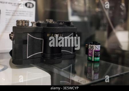 Die Vintage-Kamera steht auf einem Glasdisplay. Schwarzer Körper zeigt sichtbare mechanische Details. Filmkanister in der Nähe zeigt Rollennummer und Marke. Reflexionen A Stockfoto