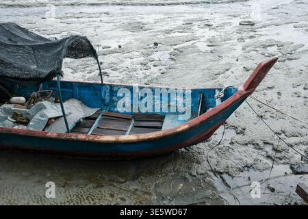 Blaues Holzboot ruht auf flachem, schlammigem Wasser. Schwarze Plane bedeckt das Innere des Bootes. Der verwitterte, rot lackierte Rumpf weist Gebrauchsspuren auf. Eisfragmente f Stockfoto