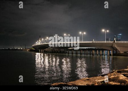 Die Nachtszene bietet eine beleuchtete Brücke über ruhiges Wasser. Licht reflektiert auf dunkler, leicht kräuselnder Oberfläche. Die Brücke überspannt einen breiten Wasserweg mit robuster Unterlage Stockfoto
