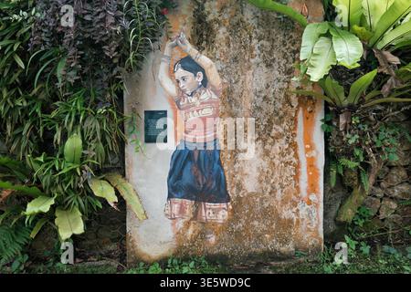 Verblasstes Wandbild zeigt eine tanzende Frau in traditioneller Kleidung. Rostflecken bedecken die Steinmauer hinter ihr. Üppig grüne Pflanzen bilden den Rahmen für die gemalte Szene. Rebe Stockfoto