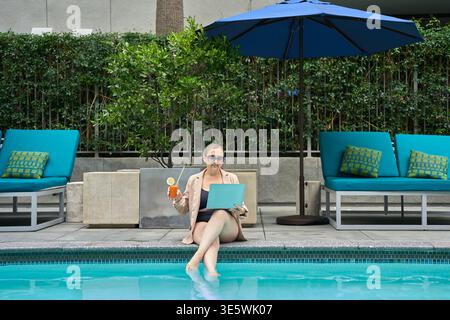 Frau in Blazer und Badeanzug sitzt am Pool mit Laptop und Cocktail. Konzept der Fernarbeit, des Lebensstils digitaler Nomaden und des Sommerurlaubs. Stockfoto