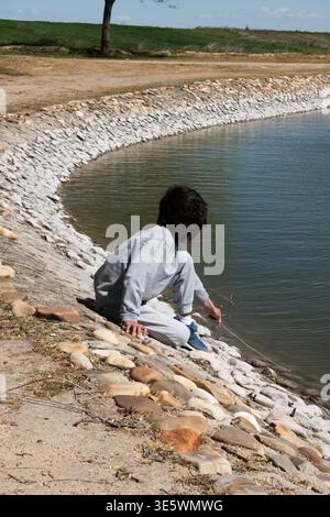 Kind beobachtet und berührt Wasser am felsigen Seeufer an einem sonnigen Tag Stockfoto