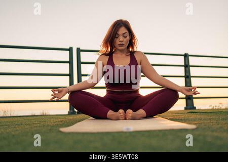 Eine Frau in sportlicher Kleidung sitzt in einer Yoga-Pose auf einer Matte vor dem Hintergrund des Sonnenuntergangs und fängt einen Moment heiterer Achtsamkeit ein. Stockfoto