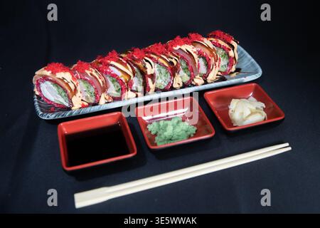 Japanische Gourmet-Sushi-Rollplatte auf einer grauen Keramikplatte, garniert mit scharfem Mayo, Drizzle und rotem Tobiko, serviert mit Sojasauce, Wasabi und Pickl Stockfoto