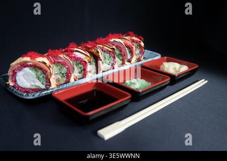 Japanische Gourmet-Sushi-Rollplatte auf einer grauen Keramikplatte, garniert mit scharfem Mayo, Drizzle und rotem Tobiko, serviert mit Sojasauce, Wasabi und Pickl Stockfoto