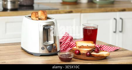Weißer Toaster, Teller mit Brotscheiben, Kirschmarmelade und Saft auf dem Tisch in der Küche Stockfoto