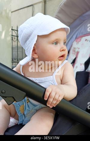 Ein Kind genießt die Natur, während es in einem Kinderwagen sitzt. Das Kind trägt einen weißen Hut und schaut sich um. Es ist ein sonniger Tag in der Stadt. Stockfoto